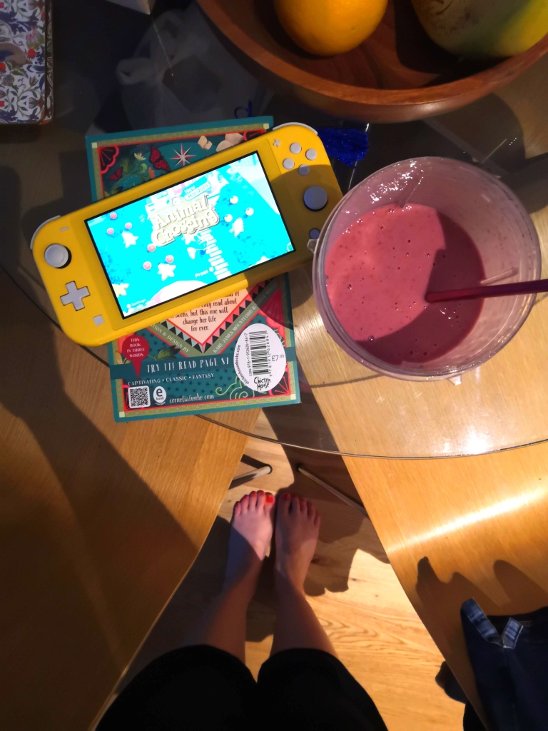A picture of a pink breakfast smoothie, a yellow nintendo switch with animal crossing playing on it and my bare feet