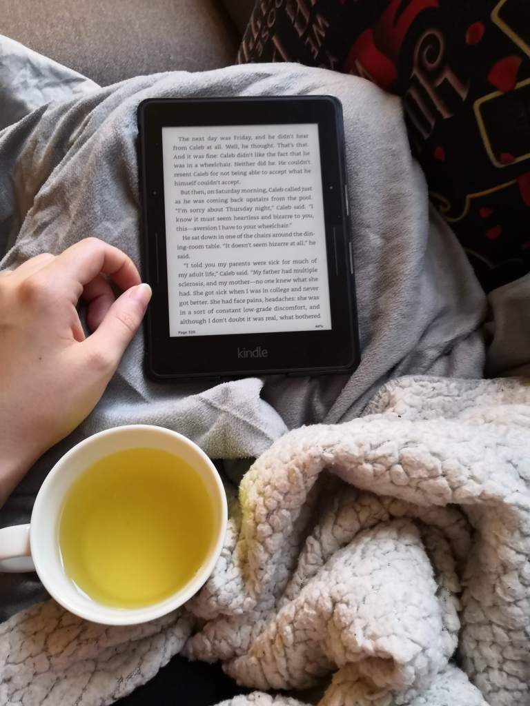 A picture of a kindle and a green tea on my leg that is wrapped up in a blanket