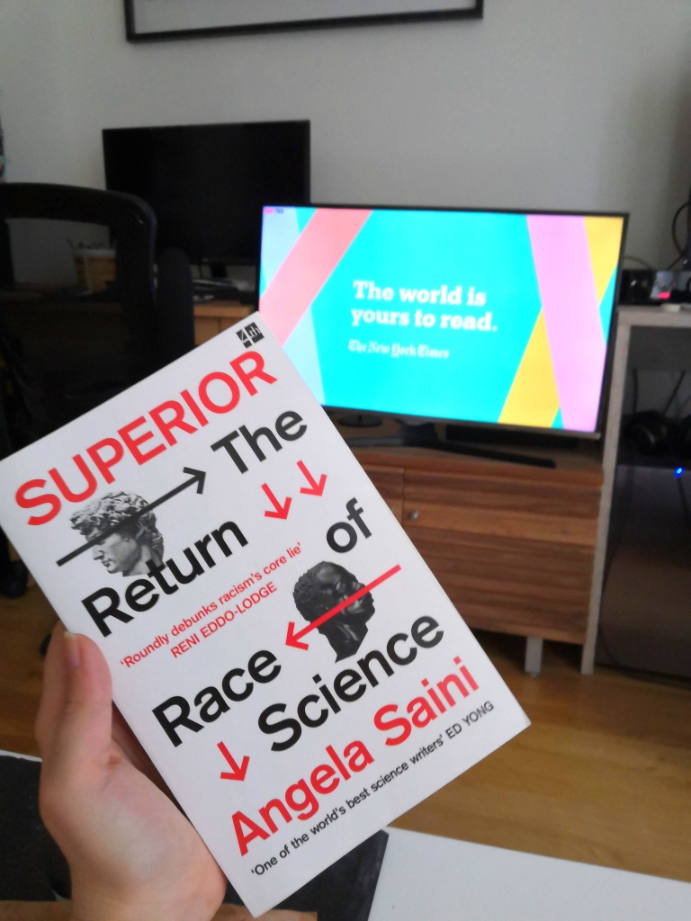 A picture of me holding a copy of Superior by Angela Saini with a TV on the background. On the TV there is a quote 'The world is yours to read'