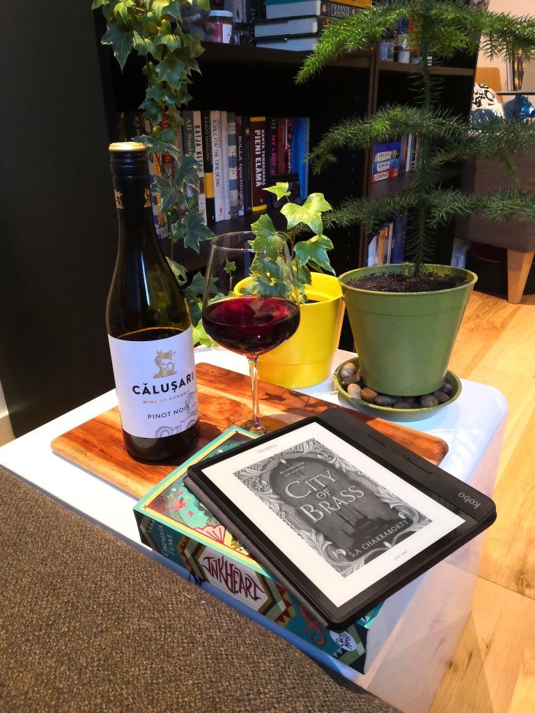 A picture taken from my couch, showing plants, a bottle of red wine and a glass full of red wine. Inkheart is underneath Kobo e-reader on a table. The e-reader has the cover of The City of Brass displayed on the screen.